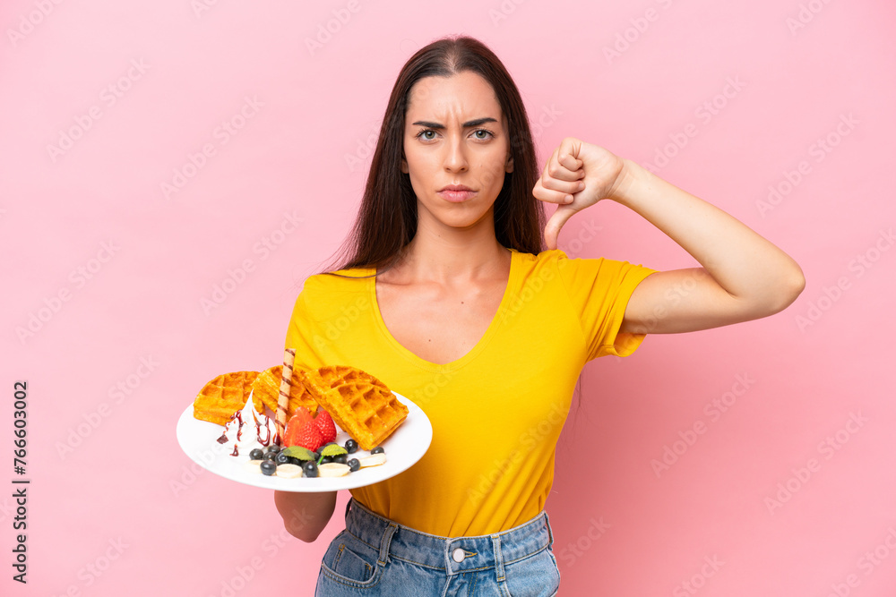 Young caucasian woman holding waffles isolated on pink background showing thumb down with negative expression