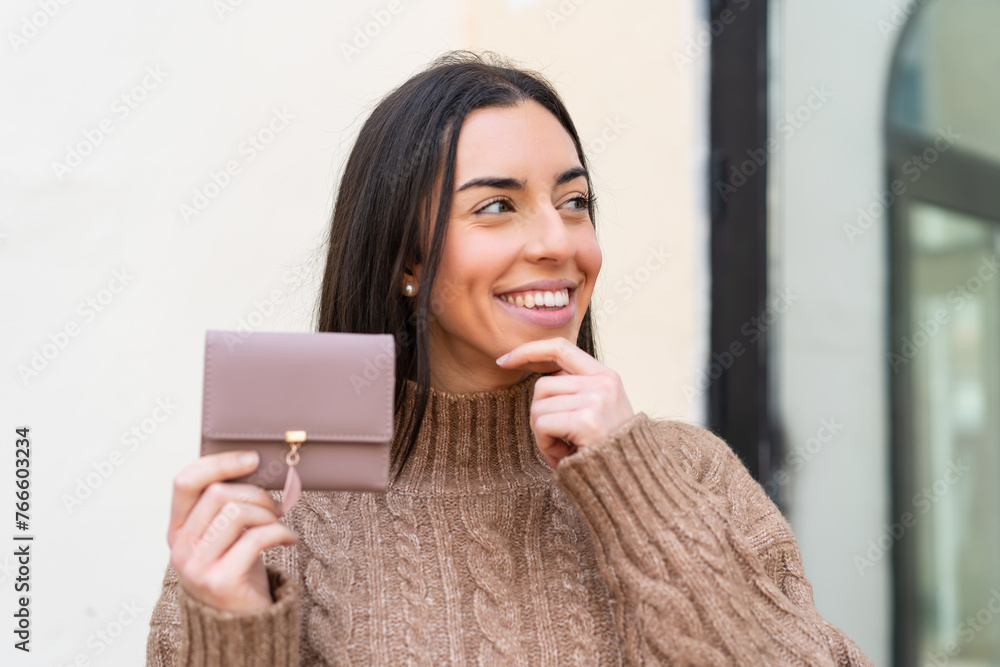 Young woman holding a wallet at outdoors thinking an idea and looking side Young woman holding a wallet at outdoors thinking an idea and looking side