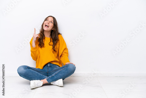 Young caucasian woman sitting on the floor isolated on white wall pointing up and surprised