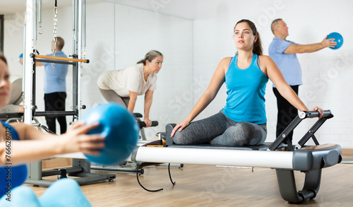 Slim young woman engaging in pilates training on reformer tower exerciser in exercise room during workout session. Persons practicing pilates with trainer