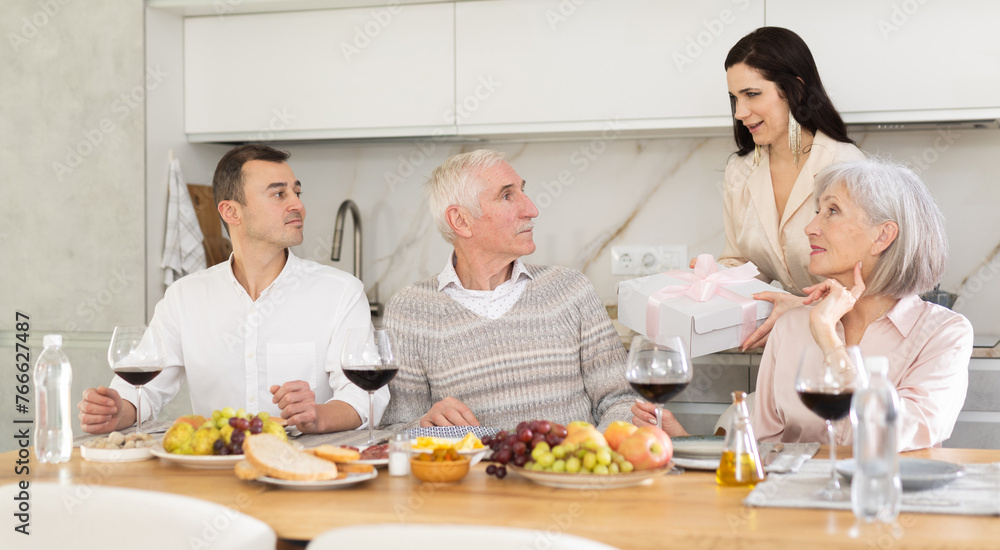 Young lady handing a gift to senior male family member sitting around table and drinking wine together at home