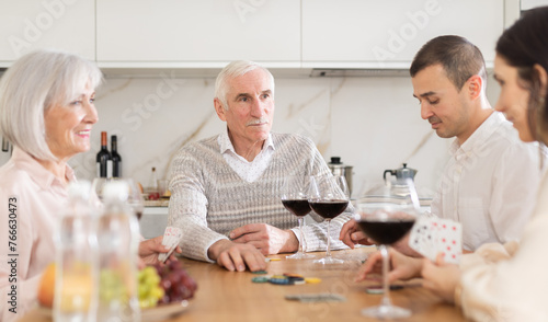 Positive elderly man spending time with family, playing poker game at dining table with wine in warm company of wife, adult son and daughter-in-law
