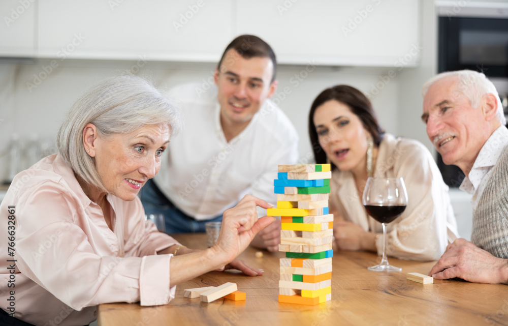 Obraz premium Carefree smiling elderly woman having fun enjoying tower building game with husband and adult children during family weekend, showcasing intergenerational bonding