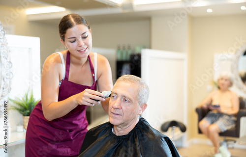 Elderly man is sitting in chair while cutting his hair in barbershop. Girl master completes creation of clients image with help of hair clipper