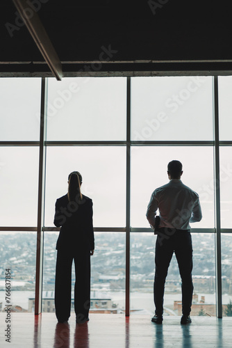 A man and a woman are standing in the office against the background of a large panoramic window. Modern spacious workspace with employees. Business partners © Дмитрий Ткачук