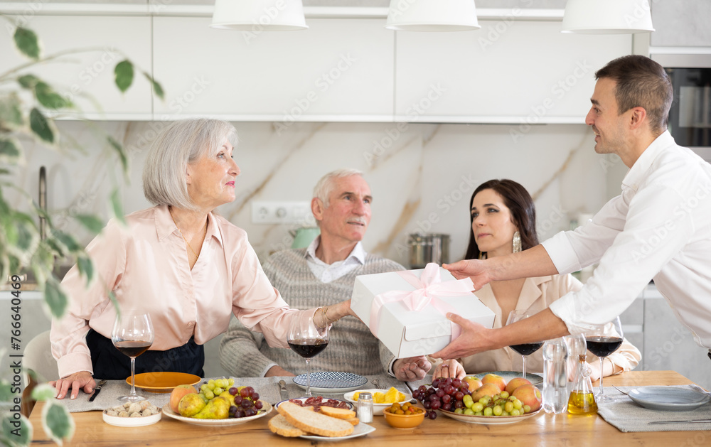 Friendly interested young man seeking to win over elderly mother of girlfriend by presenting charming gift at homely family meal in cozy kitchen interior