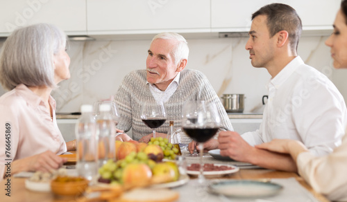 Mature spouses sitting at table, enjoying food and wine, chatting talking laughing with adult son and his girlfriend. In homely atmosphere male and female pensioner celebrate anniversary with guests