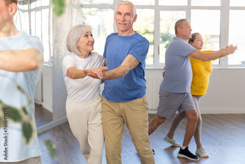 Positive senior woman and man practicing tango dance moves as a couple during a group celebration in a dance studio