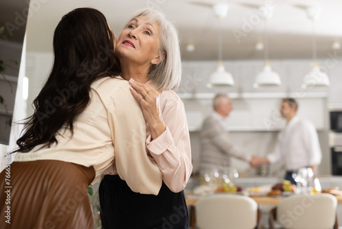 Happy elderly mother warmly welcoming with hug and kiss adult daughter coming to parents house for cozy family dinner