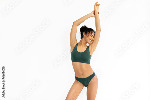 Fit latin lady standing with hands raised up, showing her ideal curves, posing in green lingerie on white studio background with copy space