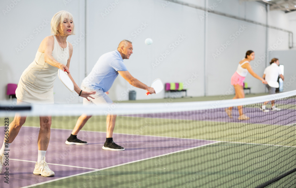 Portrait of sporty senior woman playing doubles pickleball with male partner on indoor court, ready to hit ball. Sport and active lifestyle concept