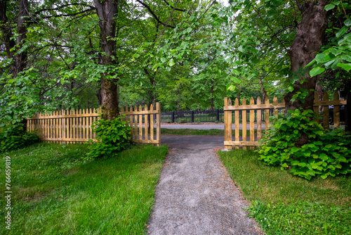 The opening of a wooden picket fence at the end of a concrete pathway. There are large mature lush green maple trees with sprigs sticking out at the bottom of the tree trunks covered in leaves