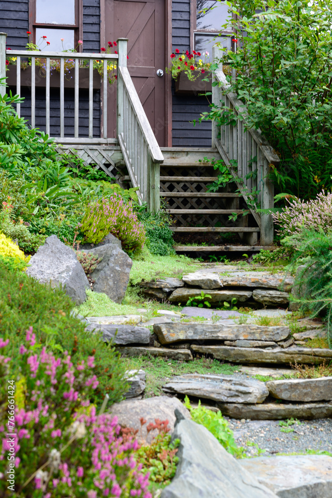 The entrance to a quaint garden shed with a wooden rail and wood steps. The brown building has a rock garden in front. The flowers are yellow, pink, and purple. The plants and trees are lush green. The entrance to a quaint garden shed with a wooden rail and wood steps. The brown building has a rock garden in front. The flowers are yellow, pink, and purple. The plants and trees are lush green.