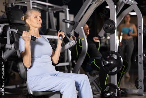 Concentrated sporty senior woman in blue activewear working out shoulders muscles on exercise machine in gym. Concept of physical activity and fitness of older adults..