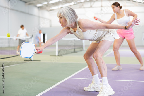 Portrait of positive fit elderly woman playing pickleball on indoor court, swinging paddle to return ball over net ..