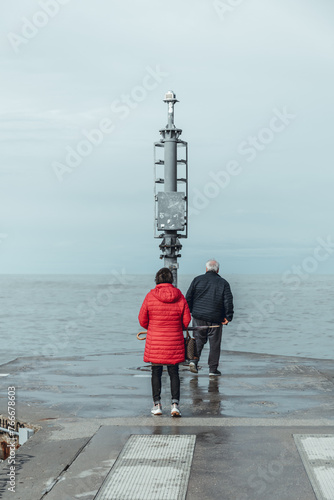 Senior couple having a romantic walk on the pier in a Riviera Romagnola beach location, bright red lady jacket, contrast with the dramatic sky in a winter afternoon. Winter sea.