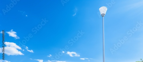 A street light pole rises from the natural landscape, with a light bulb hanging in the sky amid fluffy cumulus clouds and an electric blue atmosphere