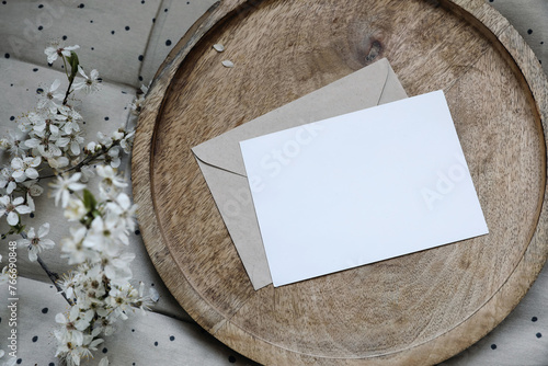Spring still life composition. Blank greeting card mockup, craft envelope. Feminine styled photo. Floral scene. Blurred white cherry tree blossoms on wooden tray. Polka dot beige seat cushion, topper.