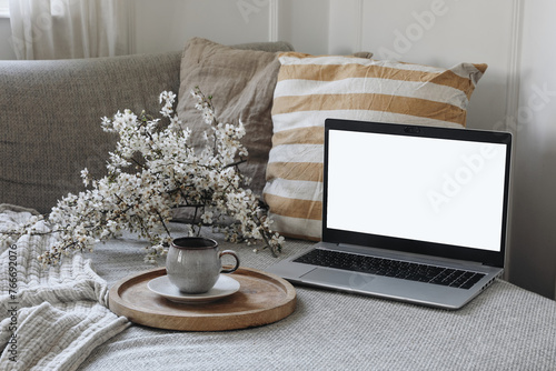 Modern spring scandinavian living room interior. Sofa with linen yellow striped cushions and cup of coffee. Cherry plum blossoms in vase. Laptop mockup with blank screen. Elegant home office decor.