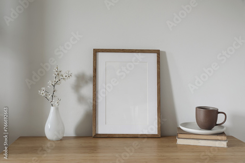 Vertical wooden picture frame, poster mockup in sunlight. Spring, easter composition. Asian interior, home office. Artistic display. Blooming cherry plum tree branches in glass vase. Wooden table.