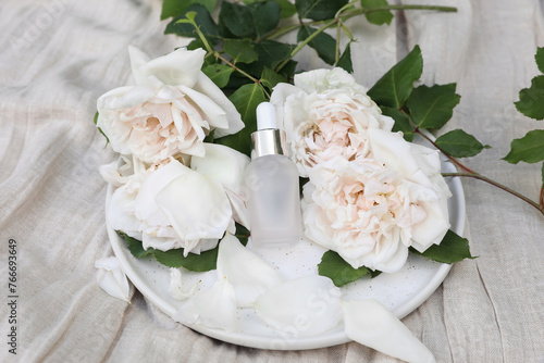 Cosmetic bottle, perfume. Rose water, essential oil. Roses flowers on beige linen table cloth background. Pink glass packaging with pipette for beauty treatment. Creative Layout. Top view, blurred