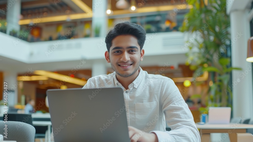 Smiling indian businessman working on laptop in modern office lobby space. Young indian student using computer remote studying, Smiling indian businessman working on laptop in modern office lobby space. Young indian student using computer remote studying,