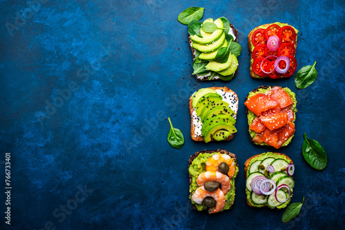 Avocado toasts with salmon, shrimp, vegetables, spinach and cream cheese, blue table background, top view
