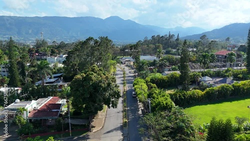Drone birds eye shot of traffic on road in Town of Dominican Republic. Green trees, grass fields and park. Silhouette of mountains in backdrop. 