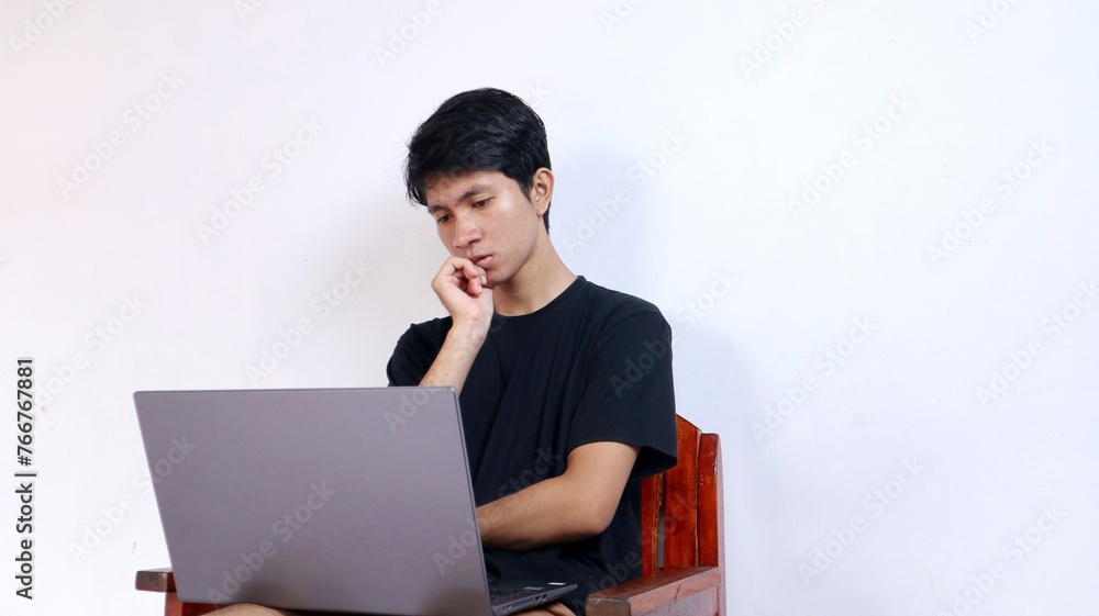 Serious young Asian man sitting doing something in front of a laptop Serious young Asian man sitting doing something in front of a laptop