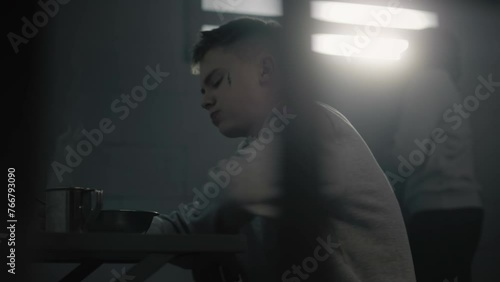 Caucasian teenage prisoner eats prison food at the table in prison cell. Young criminal serves term for crime in youth detention center or jail. Another inmate looks at the window in the background. photo