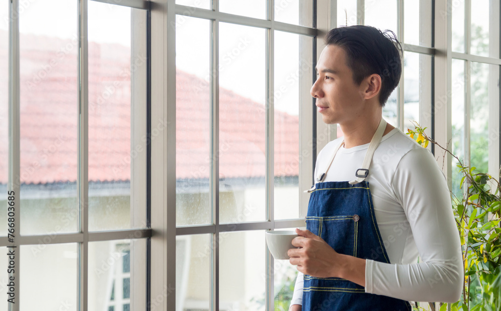 Portrait gardener adult young Asian man one person handsome standing think look and Smile relax happy hand holding drinking cup coffee in room cafe with glass window and green tree in morning time. Portrait gardener adult young Asian man one person handsome standing think look and Smile relax happy hand holding drinking cup coffee in room cafe with glass window and green tree in morning time.