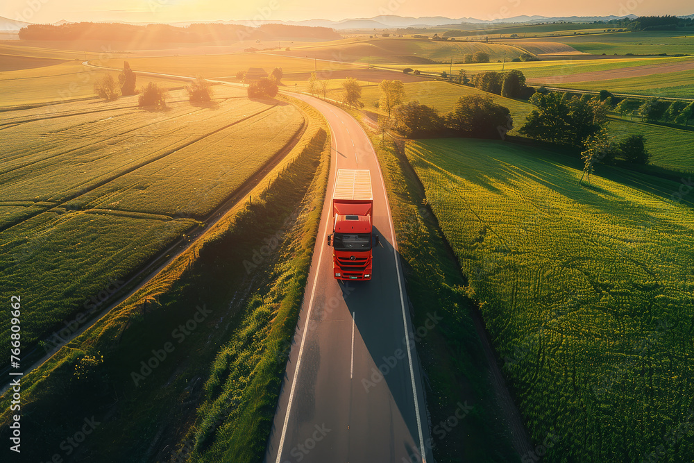 A red truck is seen driving down a road next to a beautiful, green field on a sunny day