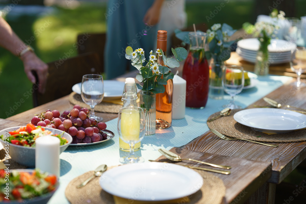 Close up shot of set table at summer garden party, grilled food. Table setting with glasses, lemonade, delicate floral and paper decoration, and bottles of summer wine.