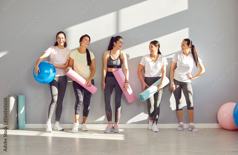 Fototapeta premium Group of young sporty girls, attractive slim happy people posing in fitness club, sportswomen team holding yoga mats. Active friends exercise or sport instruction, recreational activity together