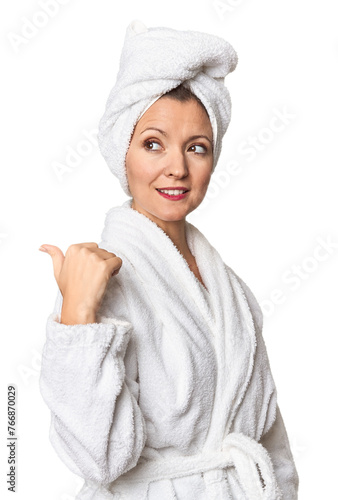 Caucasian woman in bathrobe and towel points with thumb finger away, laughing and carefree.