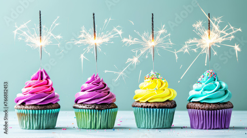 Four cupcakes with colorful frosting and lit sparklers on top are arranged festively.