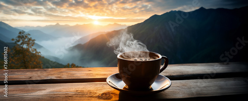 A cup of hot morning coffee with steam on a wooden table against a background of sunrise scene in the mountains. Wide scale panoramic image
