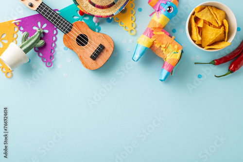 Cinco de Mayo festivities. Top view photo showcasing party essentials like petite sombrero, pinata, vihuela, cactus, bright garland of flags, spicy chilli, and nacho bowl on pastel blue background