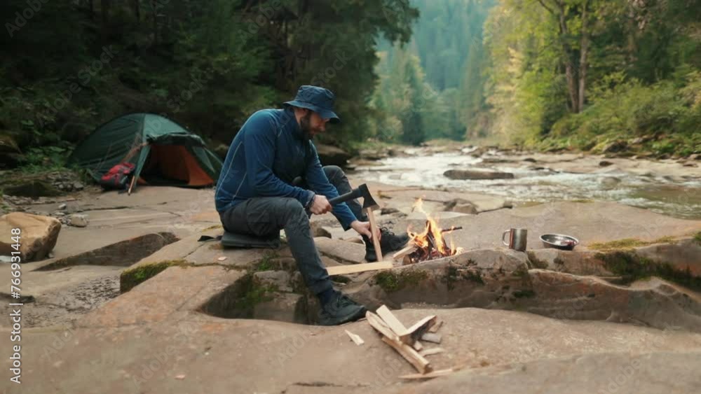 Close-up of a lone tourist sitting on a stone in a tourist camp near a fire and chopping wood Close-up of a lone tourist sitting on a stone in a tourist camp near a fire and chopping wood
