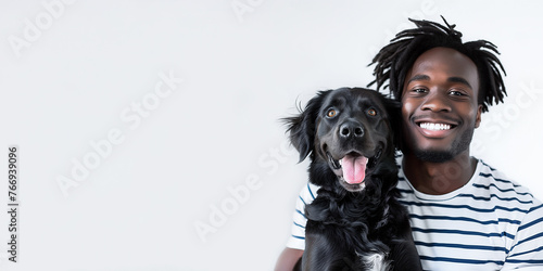 Black smiling young man with dreadlocks in a striped T-shirt hugs a black retriever dog on a white background with copy space. Human and dog friendship concept