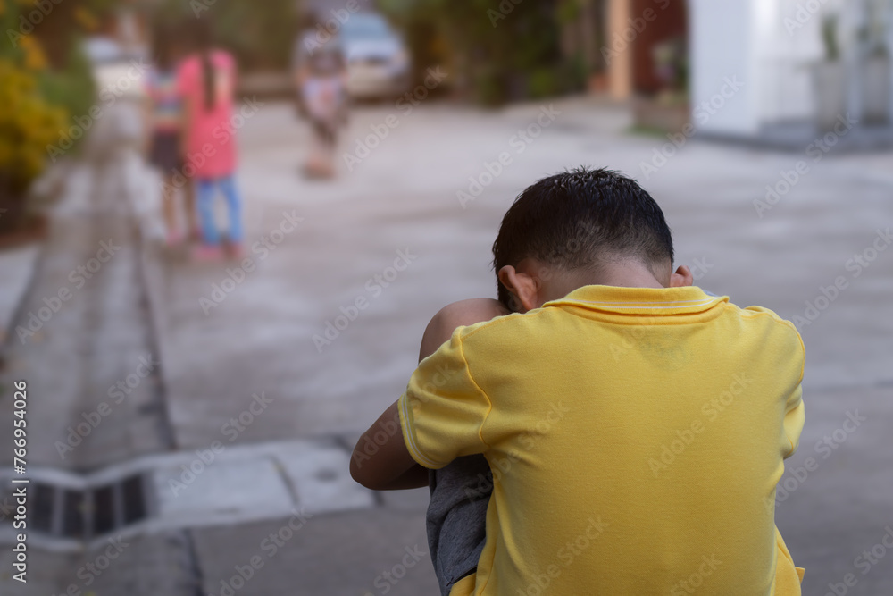 Rear side of black hair young boy sitting hugging knee on cement ground and put head down with blurry group of girls playing skateboard in front of this lonely boy. Selective focus and copy space. Rear side of black hair young boy sitting hugging knee on cement ground and put head down with blurry group of girls playing skateboard in front of this lonely boy. Selective focus and copy space.