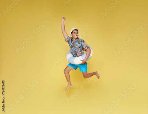 Full body young tourist asian man wear beach shirt hat hold inflatable ring jumping happy summer vacation isolated on yellow background.