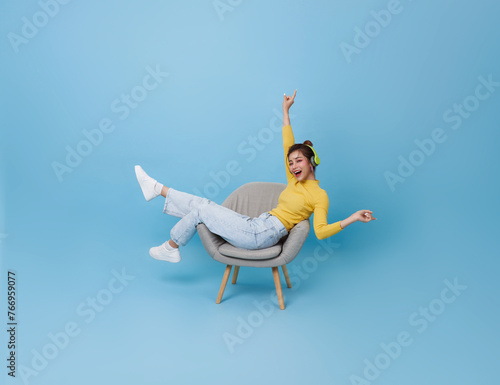 Happy young asian girl listening music with headphone dancing on chair isolated on blue background. People lifestyle concep