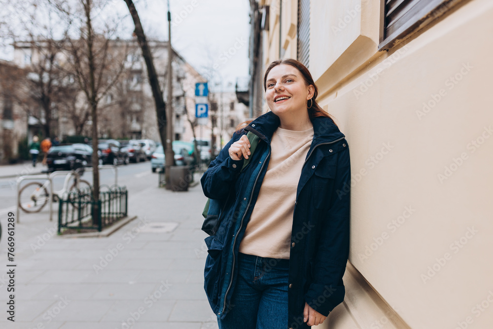 Fototapeta premium Happy young caucasian 30s woman in good mood with backpack standing on the street in the city. Urban lifestyle concept.