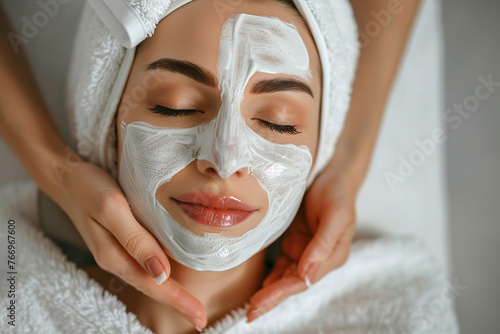 Beautiful woman Iays on the coach for beauty procedures with closed eyes wearing white robe, another hands are putting cosmetologically mask on face