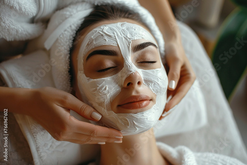 Beautiful woman Iays on the coach for beauty procedures with closed eyes wearing white robe, another hands are putting cosmetologically mask on face