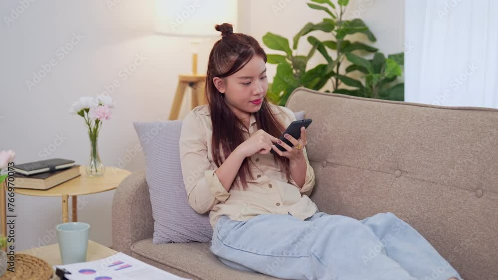 Asian businesswoman working from home, checking tasks on her smartphone on the sofa in the living room Asian businesswoman working from home, checking tasks on her smartphone on the sofa in the living room