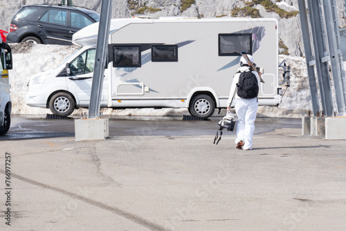 skier dressed in white overalls and skis and boots in hand walks back to her camper van in the parking lot of the ski resort