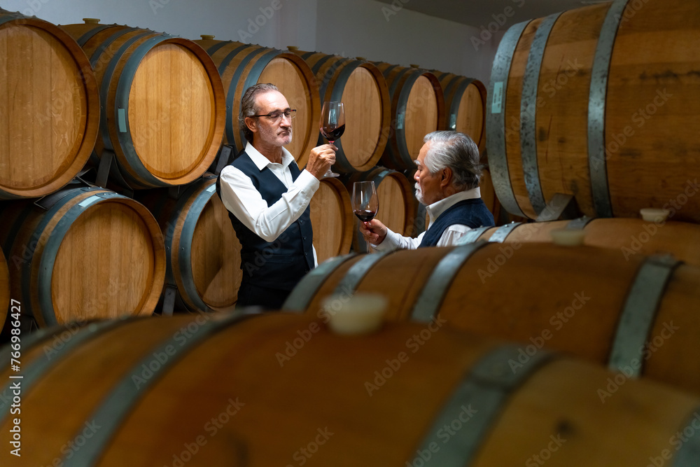Professional senior male sommelier winemaker tasting and smelling red wine in wine glass at wine cellar with wooden barrel in wine factory. Winery liquor manufacturing industry and brewery concept. Professional senior male sommelier winemaker tasting and smelling red wine in wine glass at wine cellar with wooden barrel in wine factory. Winery liquor manufacturing industry and brewery concept.
