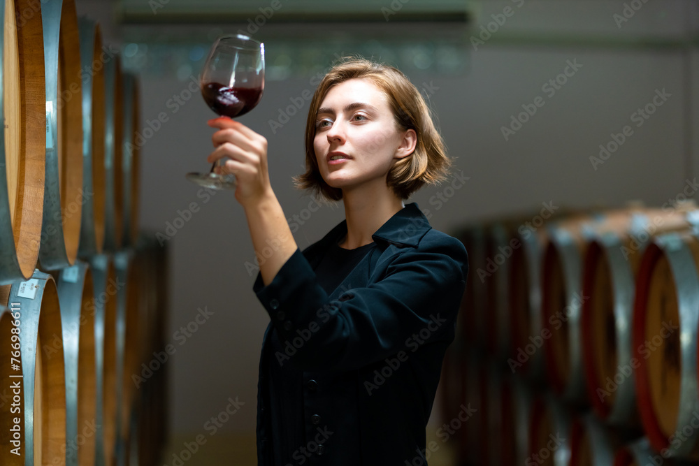 Professional Caucasian woman sommelier tasting and sniffing red wine in wine glass at wine cellar with wooden barrel. Winery brewery liquor wine factory manufacturing industry and winemaker concept.  - obrazy, fototapety, plakaty 